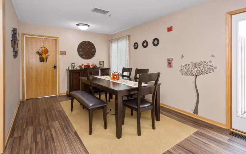 Interior dining area with a wooden table, chairs and bench on a rug, wall clock and decorative accents near a wooden door.