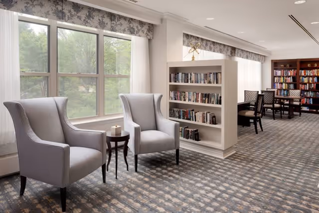 A bright and cozy reading area with two gray armchairs and a small round wooden table between them, positioned near large windows with floral valances. Behind the chairs is a white bookshelf filled with books, and further back are more bookshelves and a table with chairs, all set on a patterned carpet.