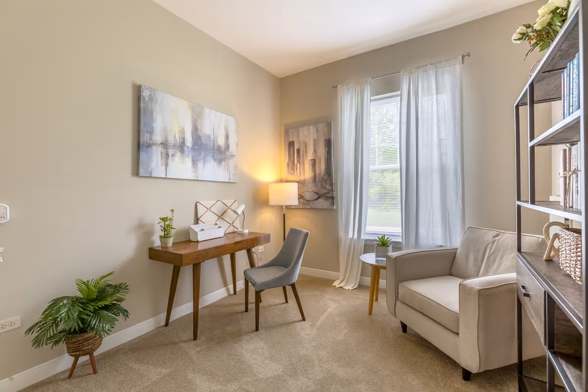 A cozy interior room with beige walls and carpet. The room features a wooden desk with a small plant, a white organizer, and a desk lamp. A gray upholstered chair is placed in front of the desk. To the right, there is a beige armchair next to a small round table with a potted plant. A tall floor lamp stands in the corner near a window with sheer white curtains. Two abstract paintings hang on the walls, and a bookshelf with books and decorative items is partially visible on the right.