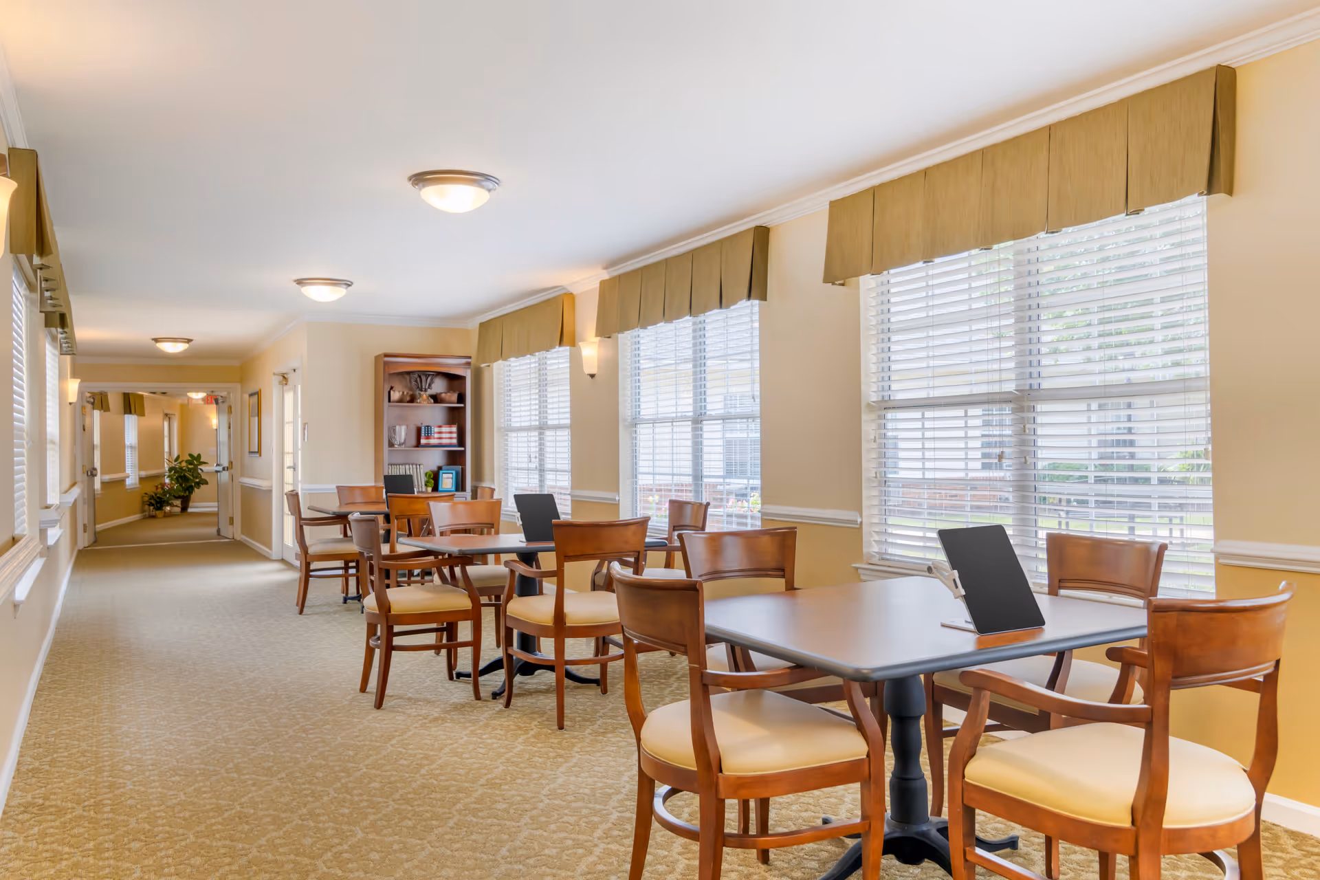 A bright hallway with large windows on the right side covered with white blinds and green valances. Several wooden tables with matching chairs are arranged along the windows, each table holding a tablet device. The hallway has beige walls and carpeted floors, with ceiling lights and a bookshelf in the background.
