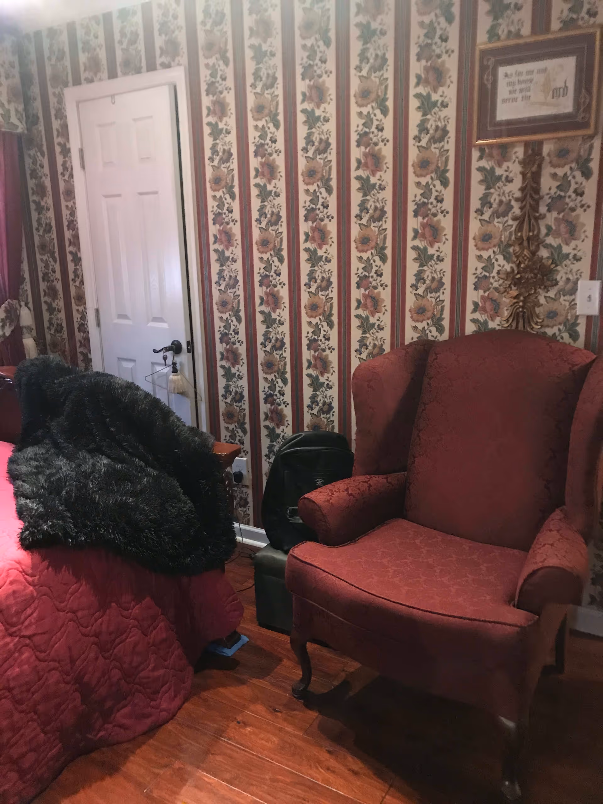 A cozy room with floral wallpaper featuring vertical stripes of flowers. There is a red upholstered armchair with a classic design on the right side and a bed with a red quilted bedspread and a dark fur blanket on the left. A white door is visible in the background, along with a framed decorative piece on the wall.