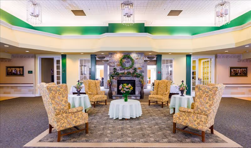 A spacious and well-lit common area with four patterned armchairs arranged around a round table covered with a white tablecloth and a vase of flowers. The room features a stone fireplace decorated with a green wreath and garland, flanked by glass-paneled doors. The ceiling has green and white accents with hanging light fixtures.