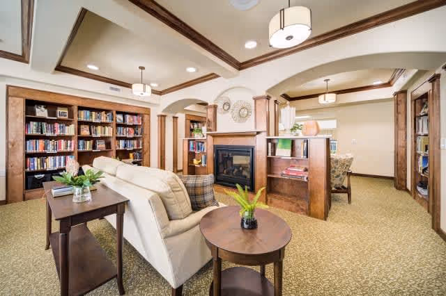 A cozy senior living facility common area with beige carpet and cream-colored walls. The room features a white sofa with plaid pillows, wooden side tables with plants, built-in wooden bookshelves filled with books, and a central fireplace with decorative wall art above it. The ceiling has recessed lighting and wooden beams, and there are archways leading to other rooms.