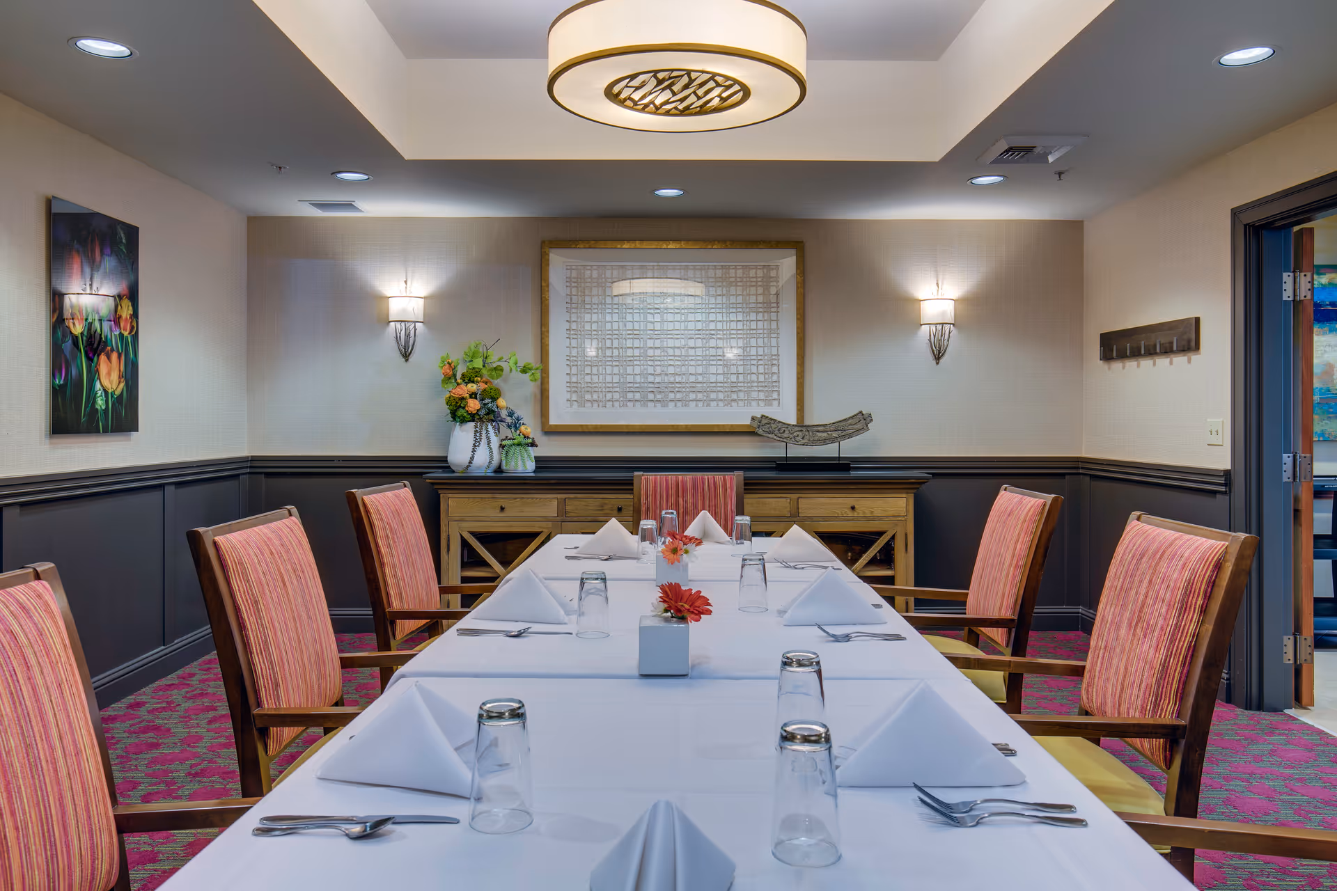 A formal dining room with a long table covered in a white tablecloth, set with folded white napkins, upside-down drinking glasses, and silverware. The chairs around the table have wooden frames and red striped upholstery. The room features a wooden sideboard with decorative items including a vase with flowers and a sculpture. The walls are decorated with framed artwork and wall sconces, and a modern circular ceiling light fixture hangs above the table.