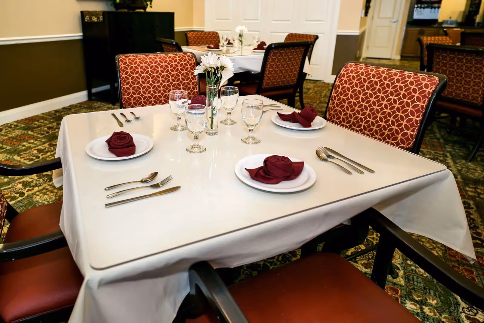 A dining table set for four with plates, folded burgundy napkins, glassware, utensils and a small flower centerpiece in a dining room.