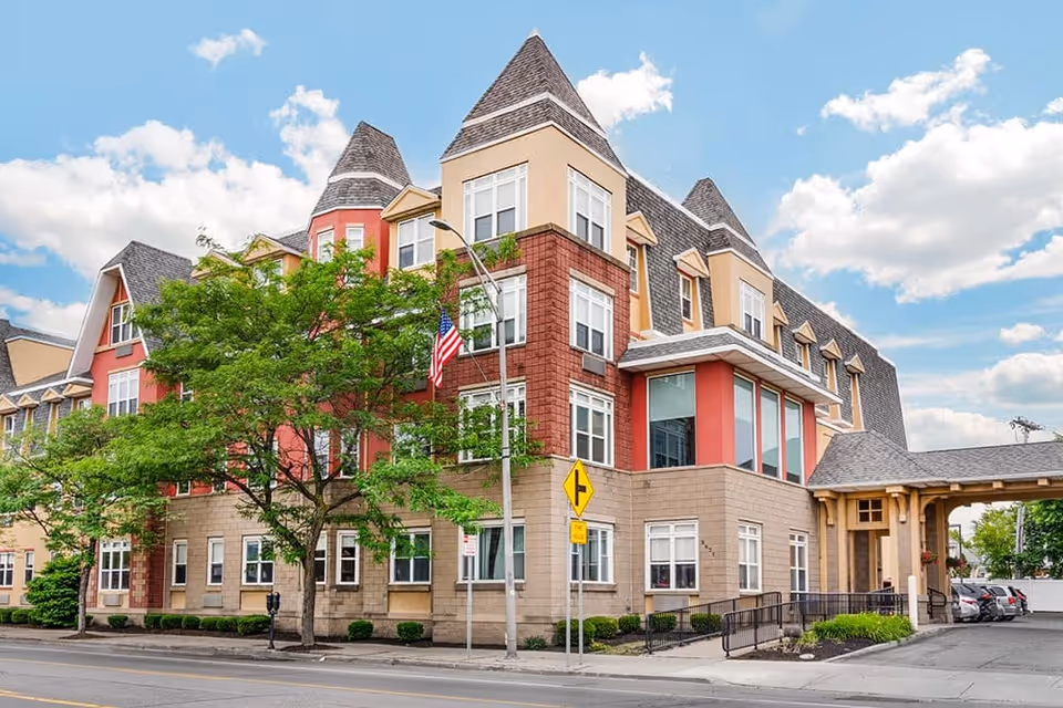 Exterior view of a multi-story senior living facility building with a mix of beige, red, and tan colors, featuring peaked roofs and multiple windows. There is a tree in front of the building, an American flag on a pole, and a street with parked cars visible on the right side under a partly cloudy sky.