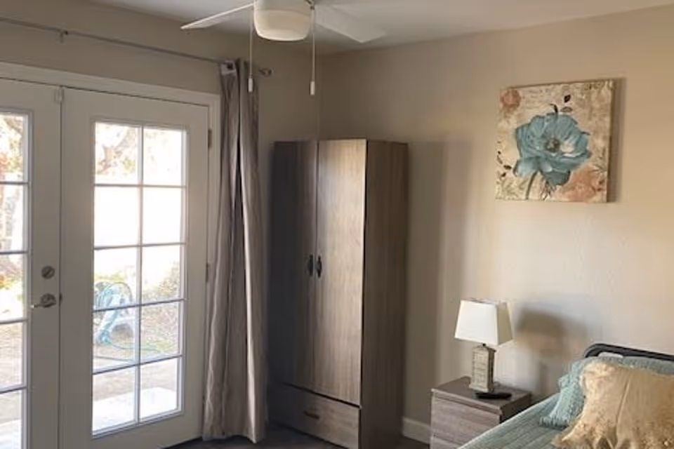 Sunlit bedroom with French doors, a wardrobe, nightstand with a lamp, and a bed with pillows.