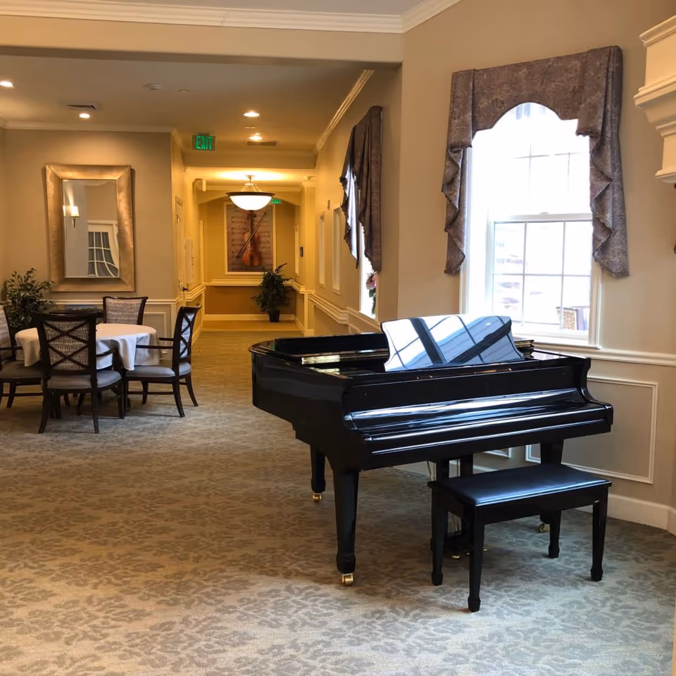 Interior view of a senior living facility hallway with a black grand piano and matching bench near a window with decorative curtains. To the left, there is a round table with four chairs and a large mirror on the wall. The hallway is carpeted and well-lit with ceiling lights and a hanging light fixture further down the corridor.