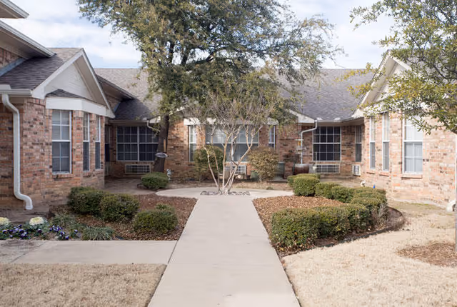 Brick single-story senior living building with a central concrete walkway leading through a landscaped courtyard with shrubs and a small tree.