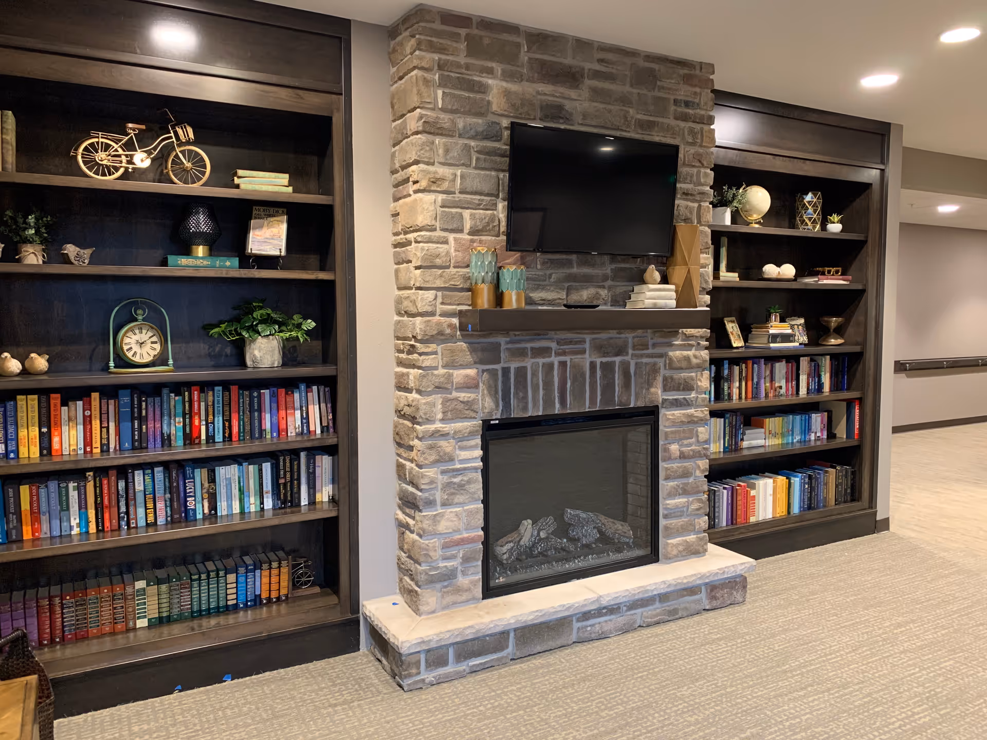 A cozy interior space featuring a stone fireplace with a mounted flat-screen TV above it. On either side of the fireplace are dark wooden bookshelves filled with books and decorative items such as plants, a clock, a globe, and small sculptures. The floor is carpeted and the walls are painted in neutral tones.