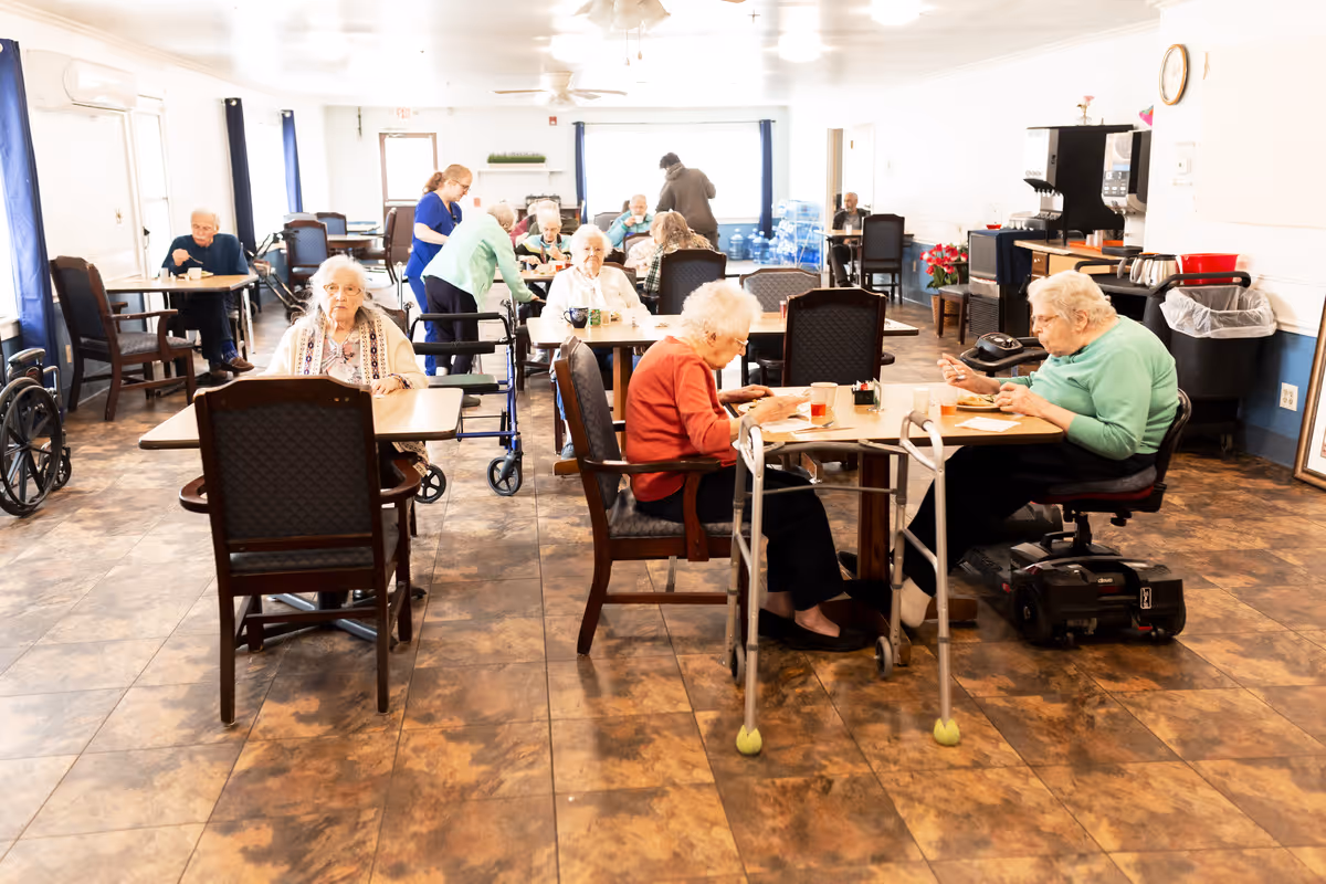 Elderly residents seated at tables eating and socializing in a bright communal dining room of a senior living facility.