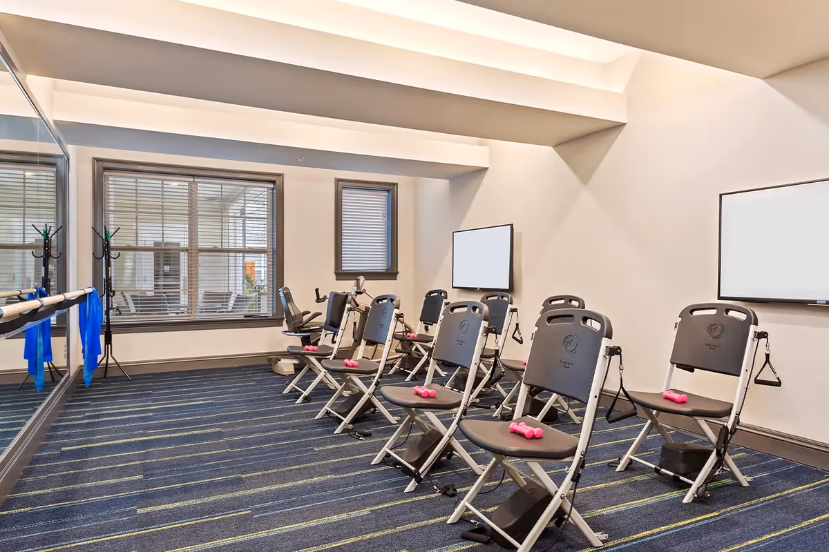 A small fitness studio with folding exercise chairs arranged in rows, pink dumbbells on the seats, wall mirrors and screens.