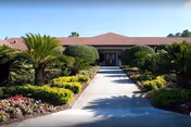 Paved walkway leading to the entrance of a single-story building surrounded by manicured landscaping and palm-like shrubs.