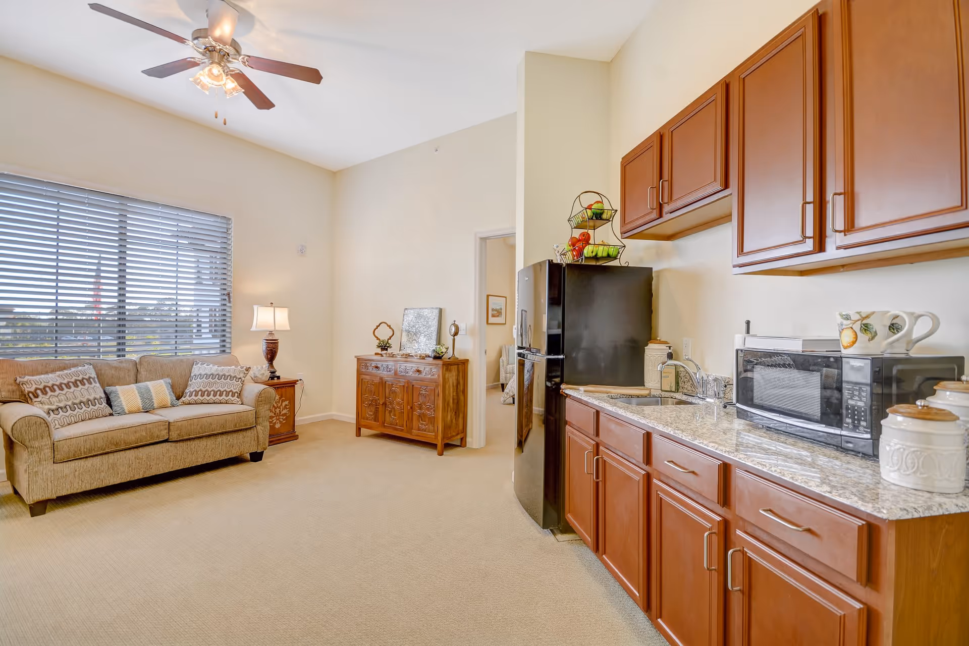 Interior view of a senior living facility room featuring a small kitchen area with wooden cabinets, a black refrigerator, a microwave, and a sink. Adjacent to the kitchen is a living area with a beige sofa, decorative pillows, a wooden side table with a lamp, and a wooden cabinet. A ceiling fan with lights is mounted on the ceiling, and a large window with blinds allows natural light into the room.