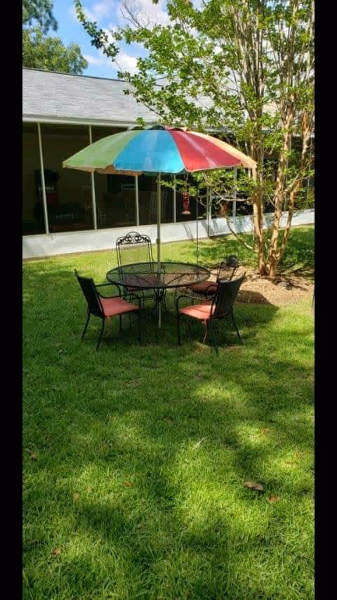 Outdoor patio area with a round metal table and four chairs with red cushions under a large multicolored umbrella on a grassy lawn. There is a tree nearby and a screened porch attached to a building in the background.