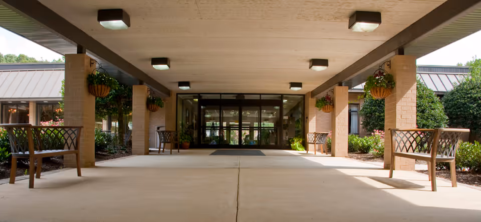 Covered entrance area of a building with benches on either side and hanging flower baskets attached to brick pillars. Glass doors lead into the building, and there are bushes and greenery around the entrance.