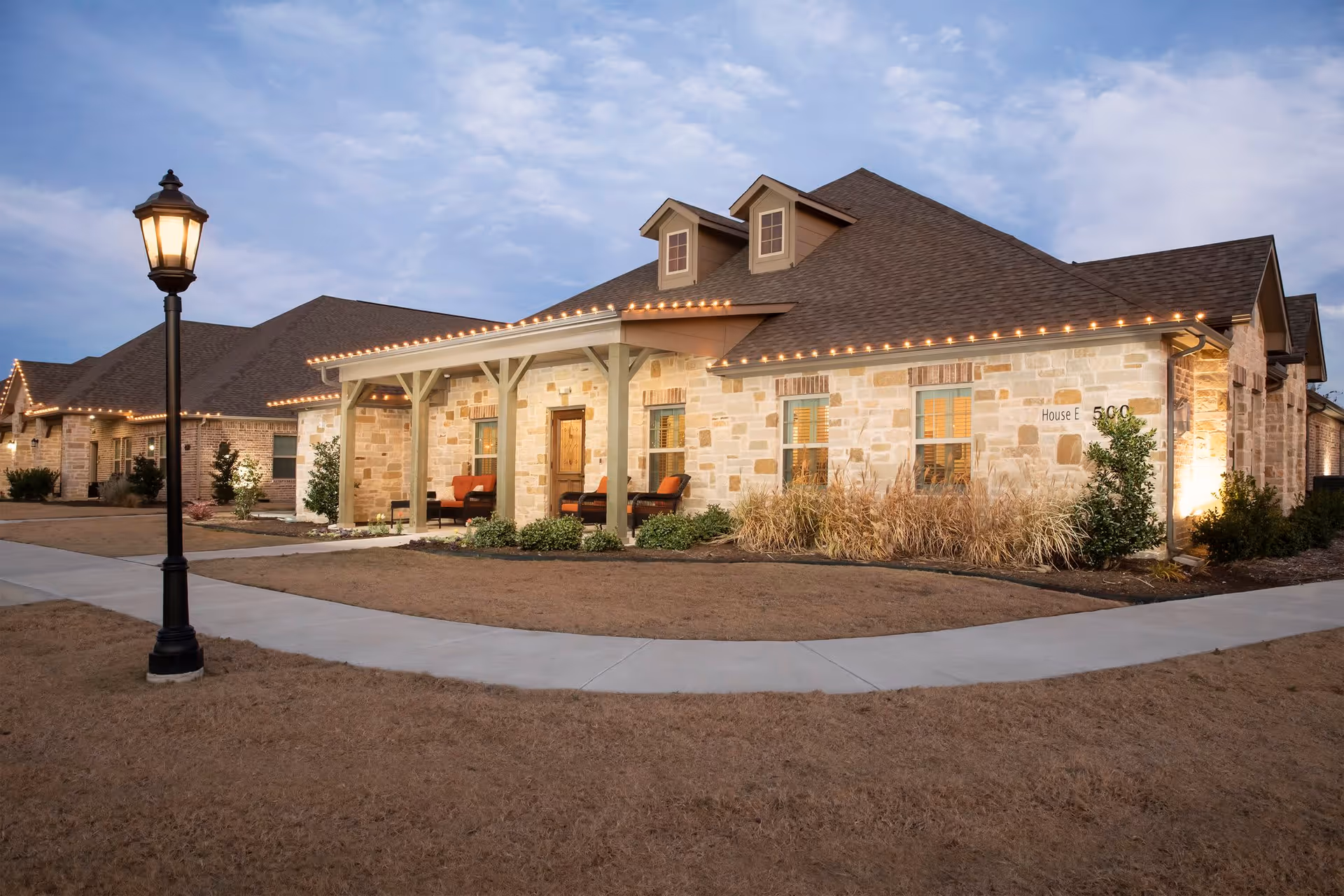 Exterior view of a single-story stone building with a covered porch featuring outdoor seating. The building has a sloped roof with dormer windows and is decorated with string lights along the eaves. A lamppost stands near a curved sidewalk in the foreground, and the surrounding area has dry grass and some landscaping plants.