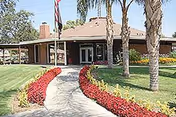 Exterior view of Country Village Apartments building with a walkway lined by red and yellow flowers leading to the entrance. There are palm trees and a flagpole with an American flag in front of the building.