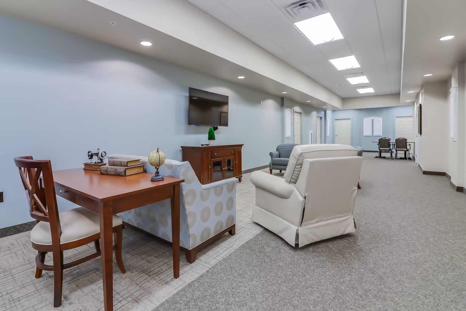 A spacious interior common area with a wooden table and chair on the left, a patterned armchair, a white recliner, and a blue armchair arranged around a wooden TV stand with a flat-screen TV mounted on the wall. The room has light blue walls, recessed ceiling lights, and a carpeted floor. In the background, there is a small table with two chairs near a light blue wall with white doors and bulletin boards.