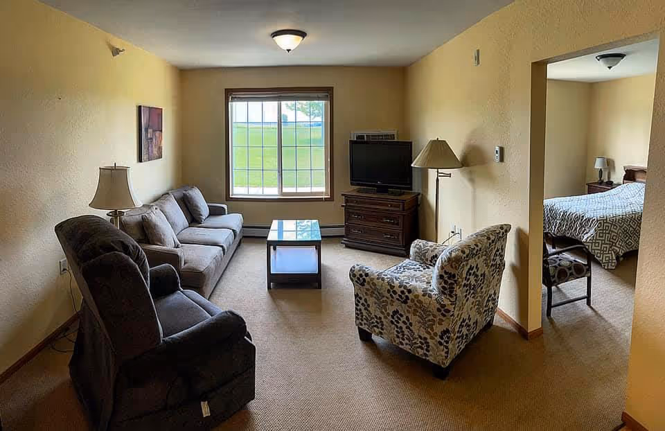A cozy living room with beige walls and carpet, featuring a gray sofa, a dark recliner chair, a patterned armchair, a glass coffee table, and two lamps. A TV sits on a wooden stand against the far wall under a window showing green grass outside. An adjacent bedroom with a bed and nightstand is visible through an open doorway.