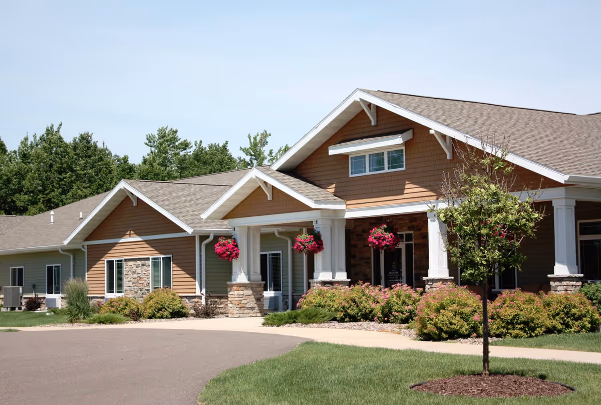 Exterior view of a single-story senior living facility building with brown siding, white trim, and stone accents. The entrance features a covered porch with white columns and hanging flower baskets. There are bushes and a small tree in the landscaped front yard under a clear blue sky.