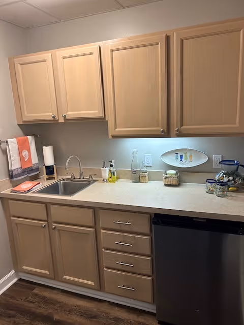A small kitchen area with light wood cabinets, a stainless steel sink, and a mini refrigerator. The countertop holds soap dispensers, a paper towel holder, a decorative plate on the wall, and some glass jars. A towel hangs on the left side near the sink.