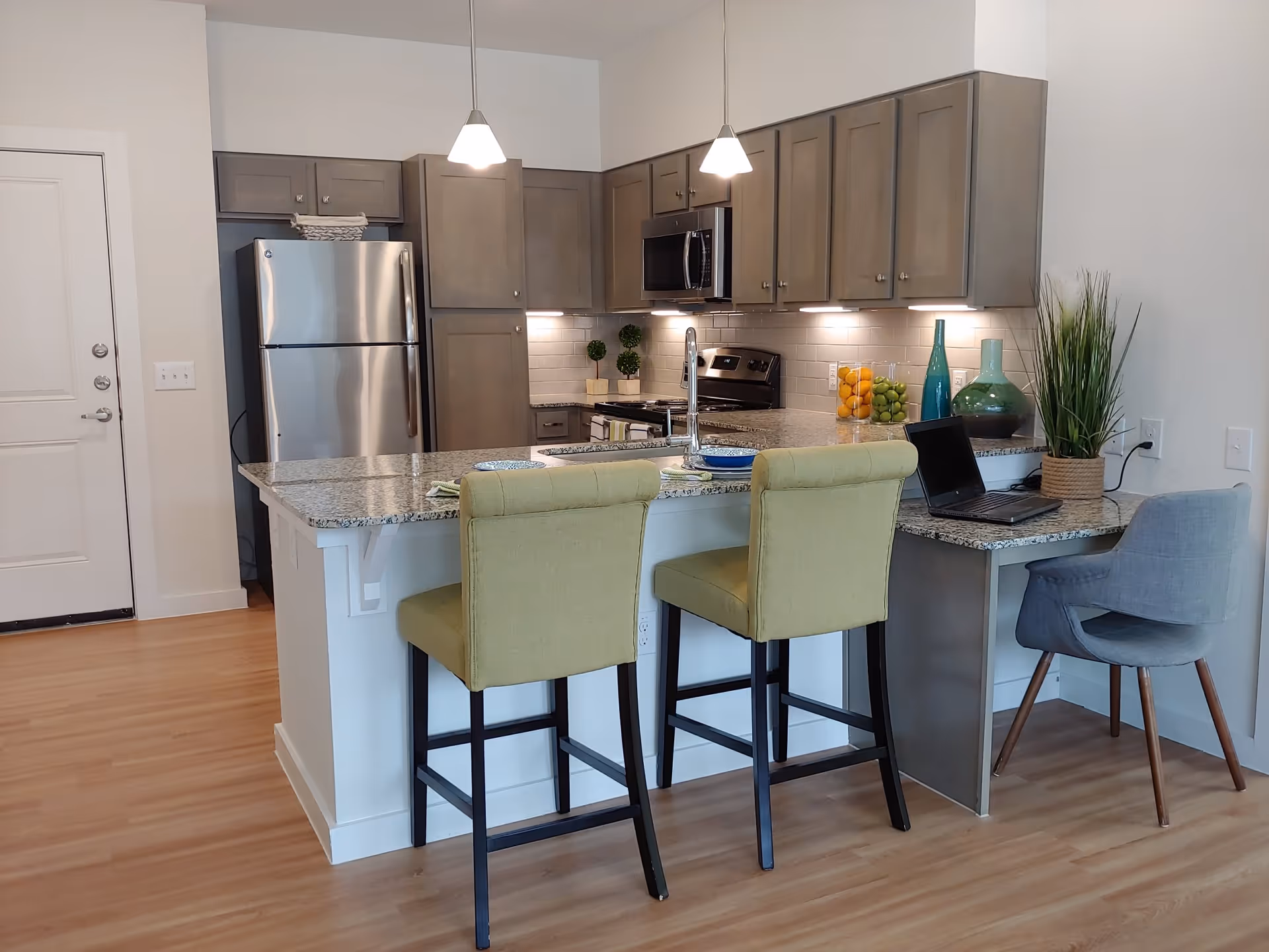 Modern kitchen area with granite countertops, two green upholstered bar stools at the kitchen island, and a gray chair at a small desk area. The kitchen features stainless steel appliances including a refrigerator, microwave, and stove, with gray cabinets and under-cabinet lighting. Decorative items include a potted plant, glass jars with fruit, and vases on the countertop.