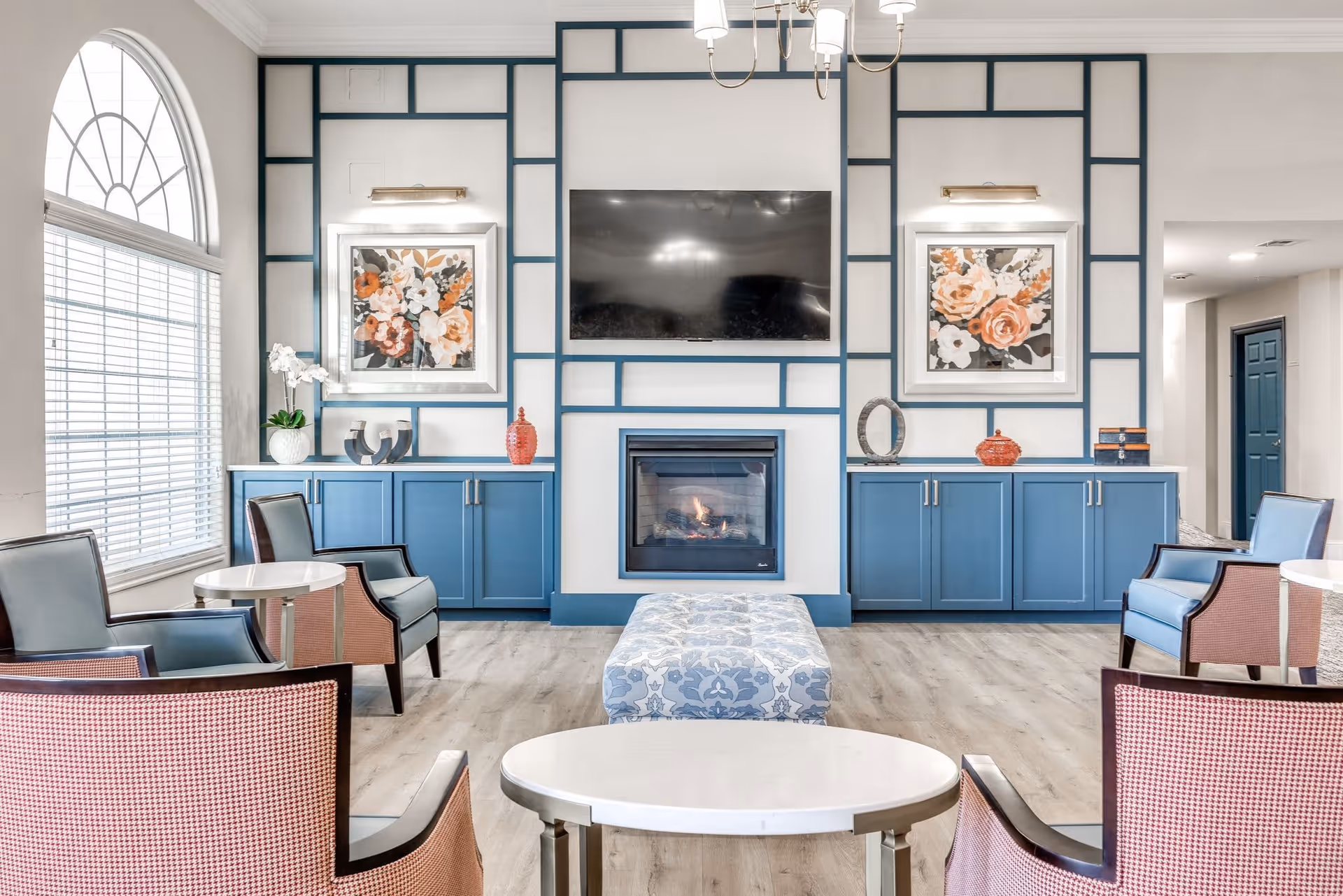 A bright and elegant senior living facility common area with blue cabinetry, a wall-mounted flat screen TV above a modern fireplace, floral artwork on the walls, and comfortable seating including armchairs and a patterned ottoman. Large arched window with blinds allows natural light to fill the room.