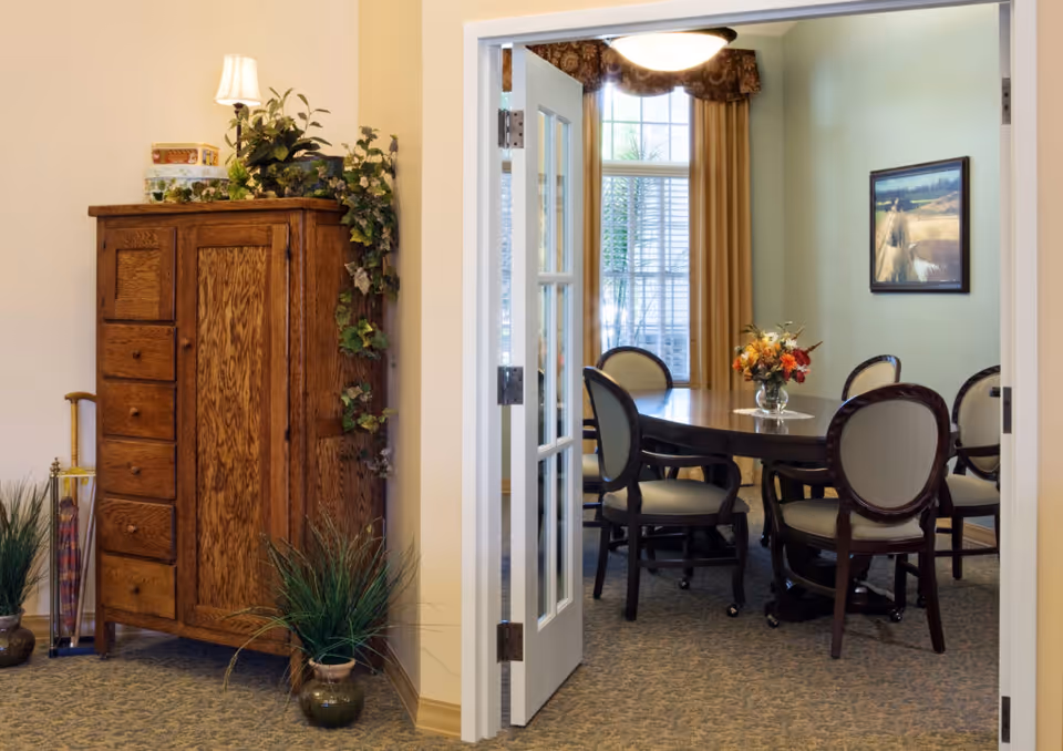 View through open double doors into a dining room with a round wooden table surrounded by six upholstered chairs. A vase with a colorful flower arrangement is on the table. The room has a large window with beige curtains and a framed landscape painting on the wall. In the foreground, there is a wooden cabinet with drawers and a lamp on top, along with some green plants on the floor and climbing the cabinet.