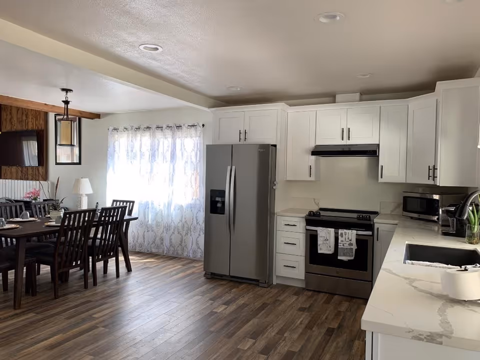 Open kitchen and dining area with white cabinets, stainless steel appliances, marble-look countertops, and a wooden dining table.