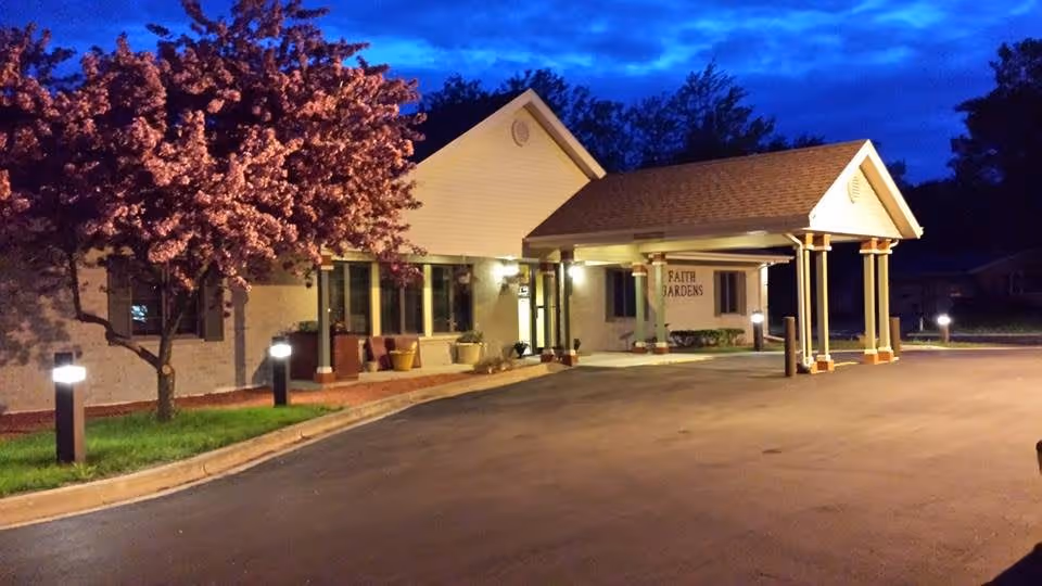 Exterior view of Faith Gardens Memory Care facility at dusk, showing a well-lit entrance with a covered drop-off area, a blooming tree with pink flowers on the left, and a paved driveway.
