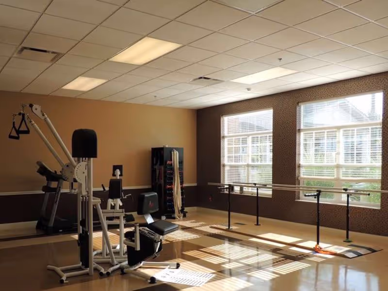 A well-lit exercise room with large windows allowing natural light to enter. The room contains various exercise equipment including a weight machine, a treadmill, and parallel bars for walking or balance exercises. The walls are painted in warm tones with a patterned wallpaper on one side.