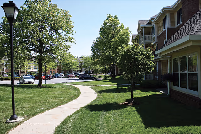 A curved concrete walkway runs through a grassy area with trees and a lamppost, leading towards a parking lot with several cars. On the right side, there is a multi-story brick building with large windows and balconies. The sky is clear and blue.