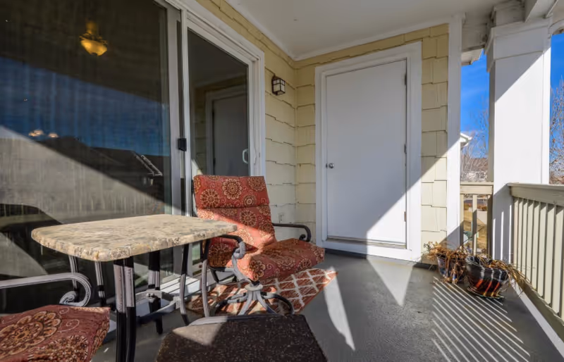 A small outdoor balcony area with two cushioned chairs featuring red patterned upholstery, a small square table with a stone-like surface, a white door, and a sliding glass door. There are two potted plants on the floor near the railing, and sunlight casts shadows across the space.