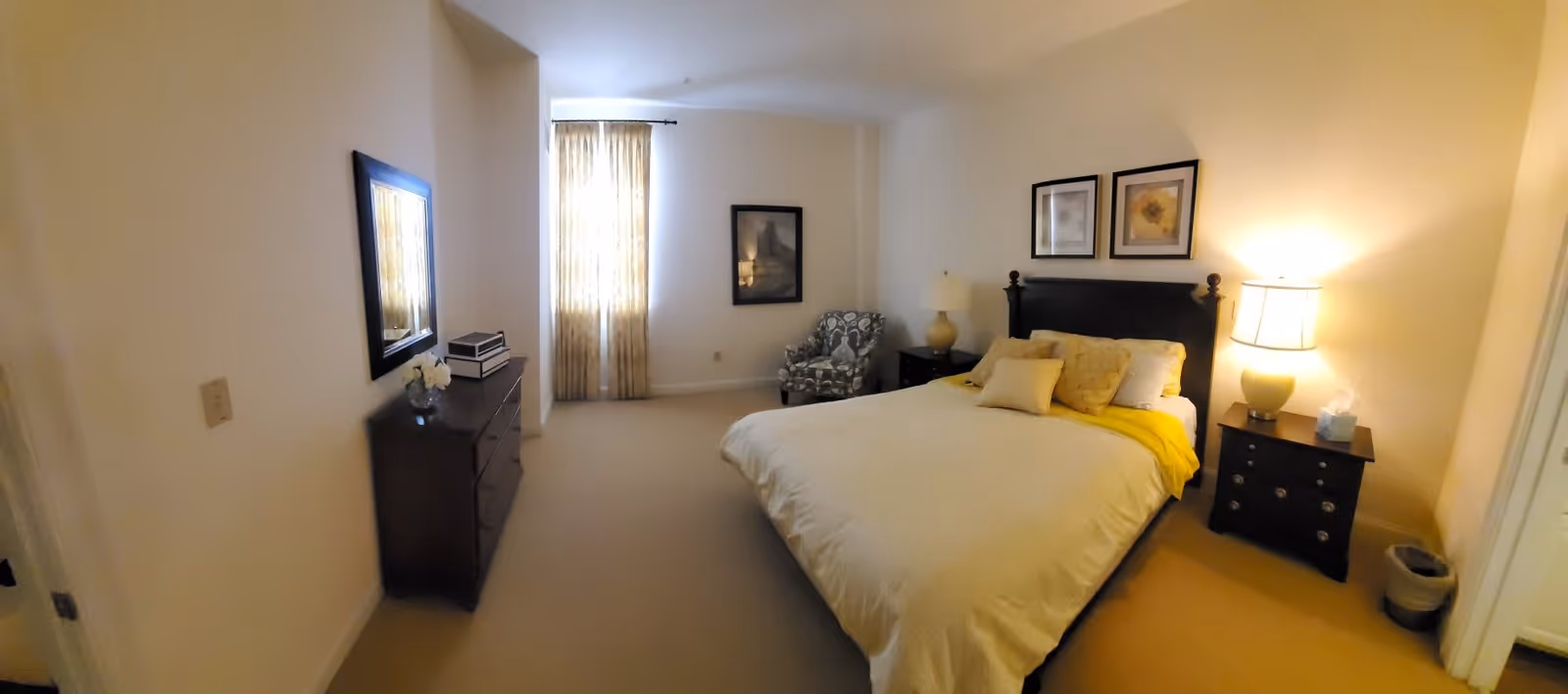 A bedroom with a large bed covered in white bedding and yellow accents, two nightstands with lamps on either side of the bed, a patterned armchair near a window with curtains, a dresser with a mirror above it, and framed artwork on the walls.