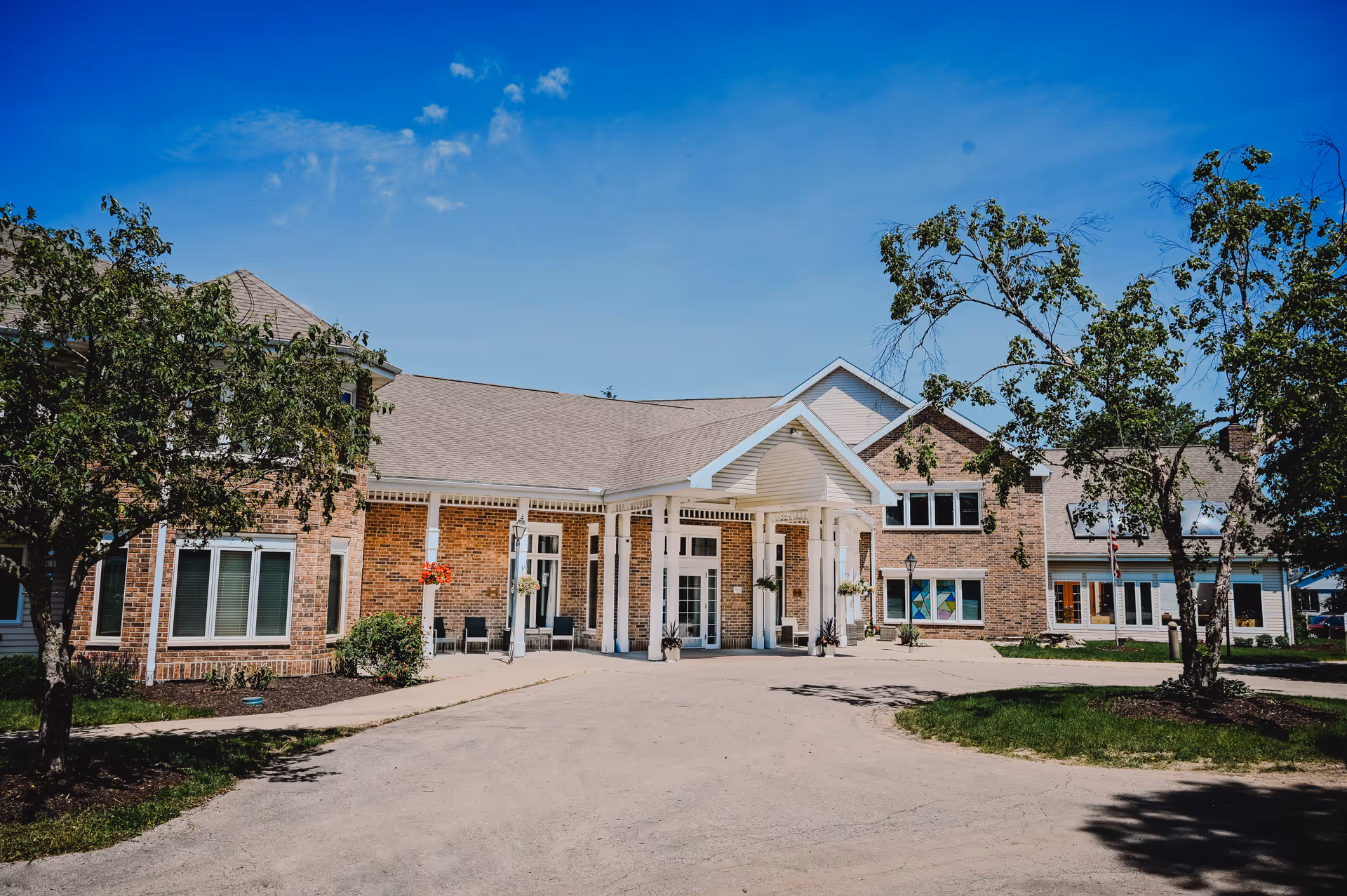 Exterior view of a senior living facility named Eden Vista Madison, showing a large brick building with white trim, multiple windows, a covered entrance with white columns, and surrounding trees under a clear blue sky.