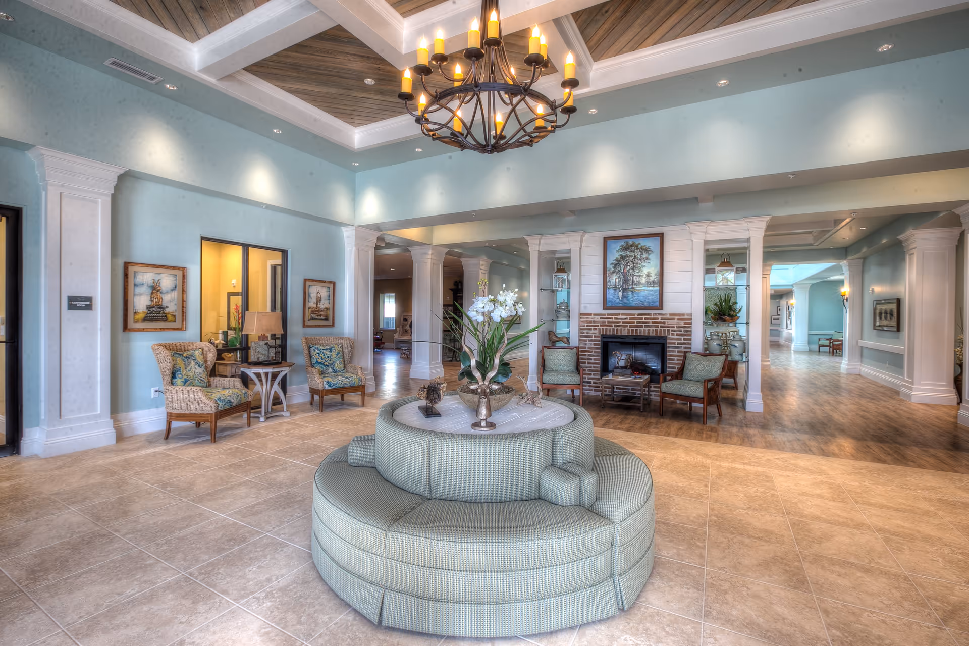 A spacious and elegantly decorated common area in a senior living facility with light blue walls, a large circular cushioned seating arrangement in the center with a floral arrangement on top, wicker and wooden chairs with patterned cushions, a brick fireplace with a painting above it, and a chandelier hanging from a wooden coffered ceiling.