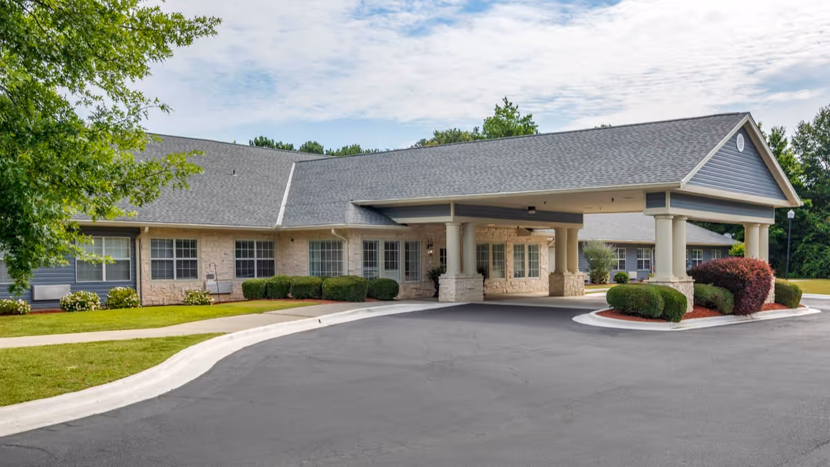 Front entrance of a one-story senior living facility with a covered porte-cochère, landscaped shrubs, and a paved driveway.