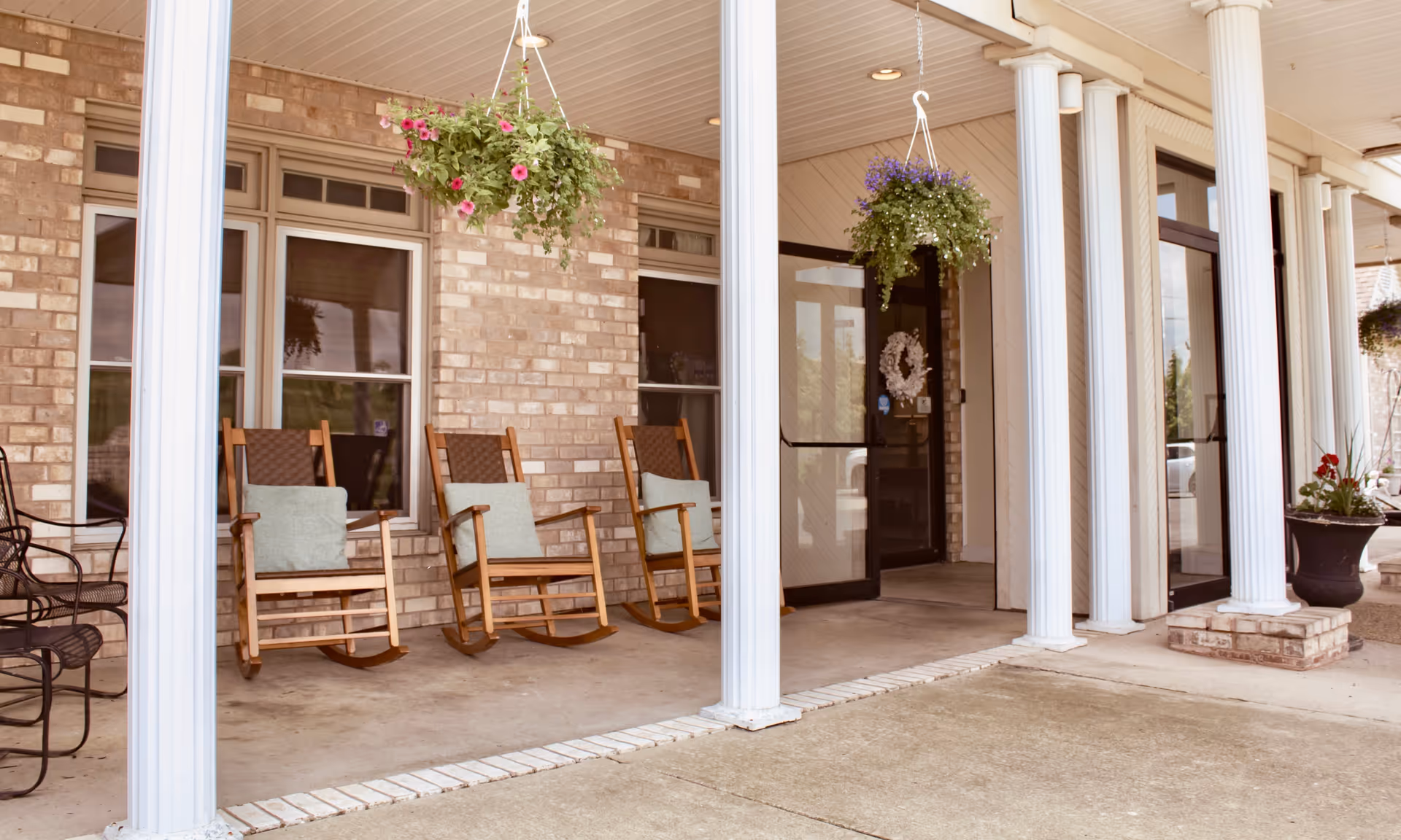 Front porch area of a facility with brick walls, white columns, hanging flower baskets, wooden rocking chairs with cushions, and a glass door entrance decorated with a wreath.