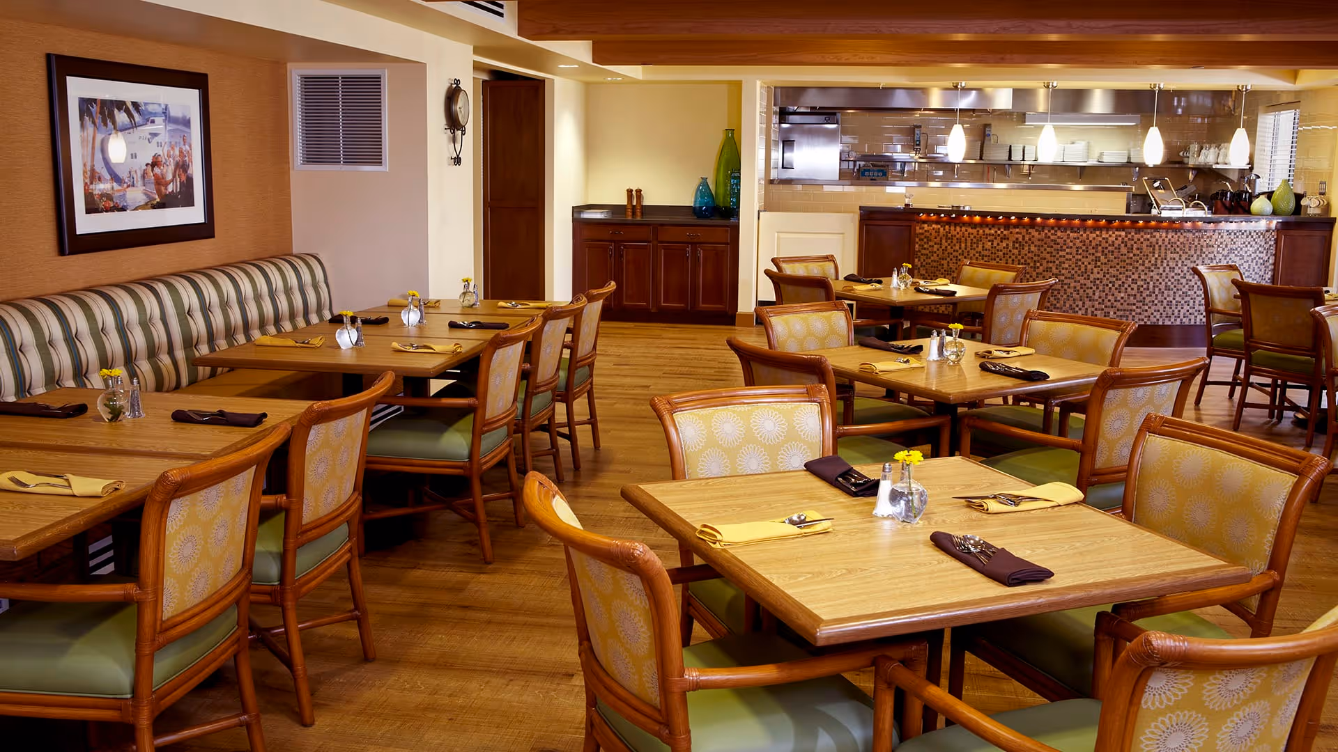 A dining room with multiple wooden tables and chairs arranged neatly. Each table is set with yellow and brown napkins, silverware, and small glass vases containing yellow flowers. There is a striped cushioned bench along one wall, a framed painting, and a serving counter with kitchen equipment visible in the background.