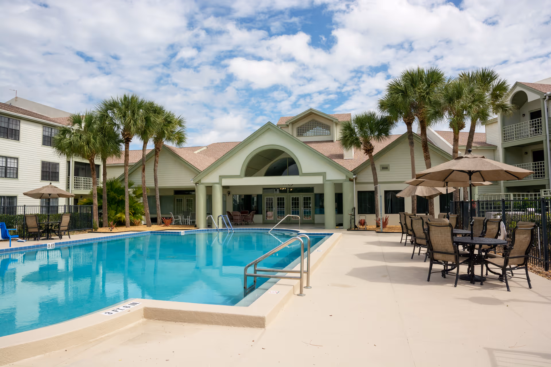 Outdoor swimming pool area at Cascade Heights with lounge chairs, tables with umbrellas, palm trees, and a building with a covered patio in the background under a partly cloudy sky.