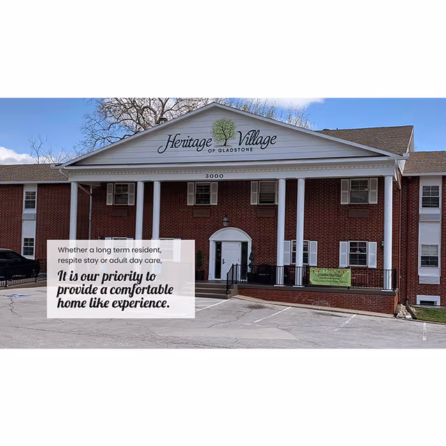 Front exterior view of Heritage Village of Gladstone, a two-story brick building with white columns and a sign displaying the facility's name and address number 3000. There is a parking lot in front and a banner on the right side of the building.