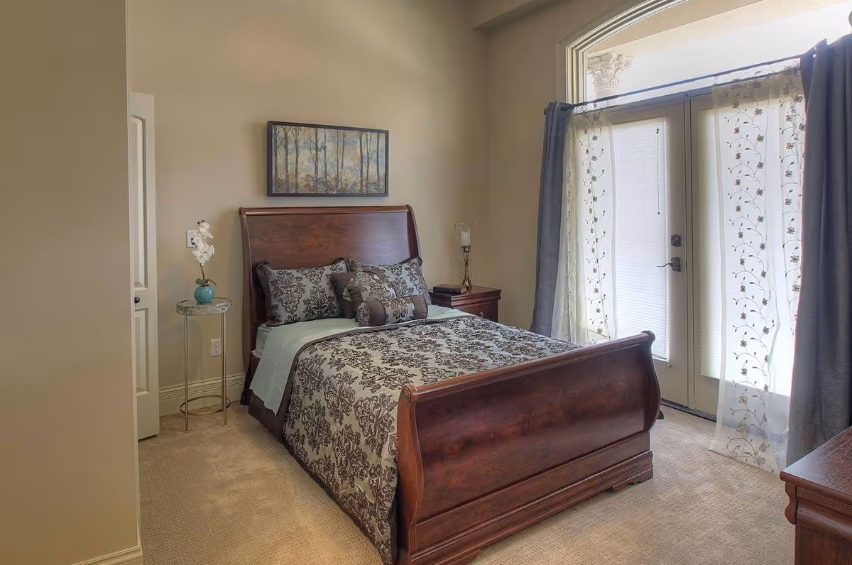 Sunlit bedroom with a wooden sleigh bed, patterned bedding, nightstands and French doors with sheer curtains.