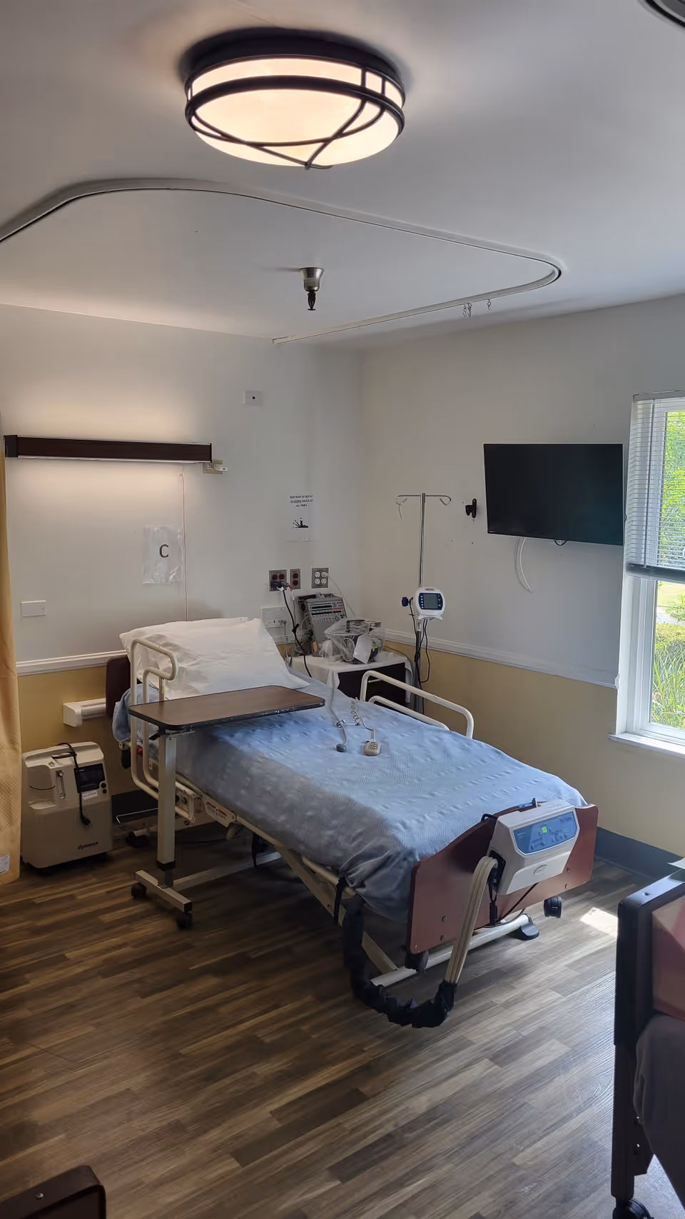 A hospital-style patient bed with blue bedding in a care facility room. The bed has side rails and an overbed table. Medical equipment, including an IV stand and monitoring devices, are positioned near the bed. A flat-screen TV is mounted on the wall next to a window with blinds. The room has wood-patterned flooring and a ceiling light fixture.