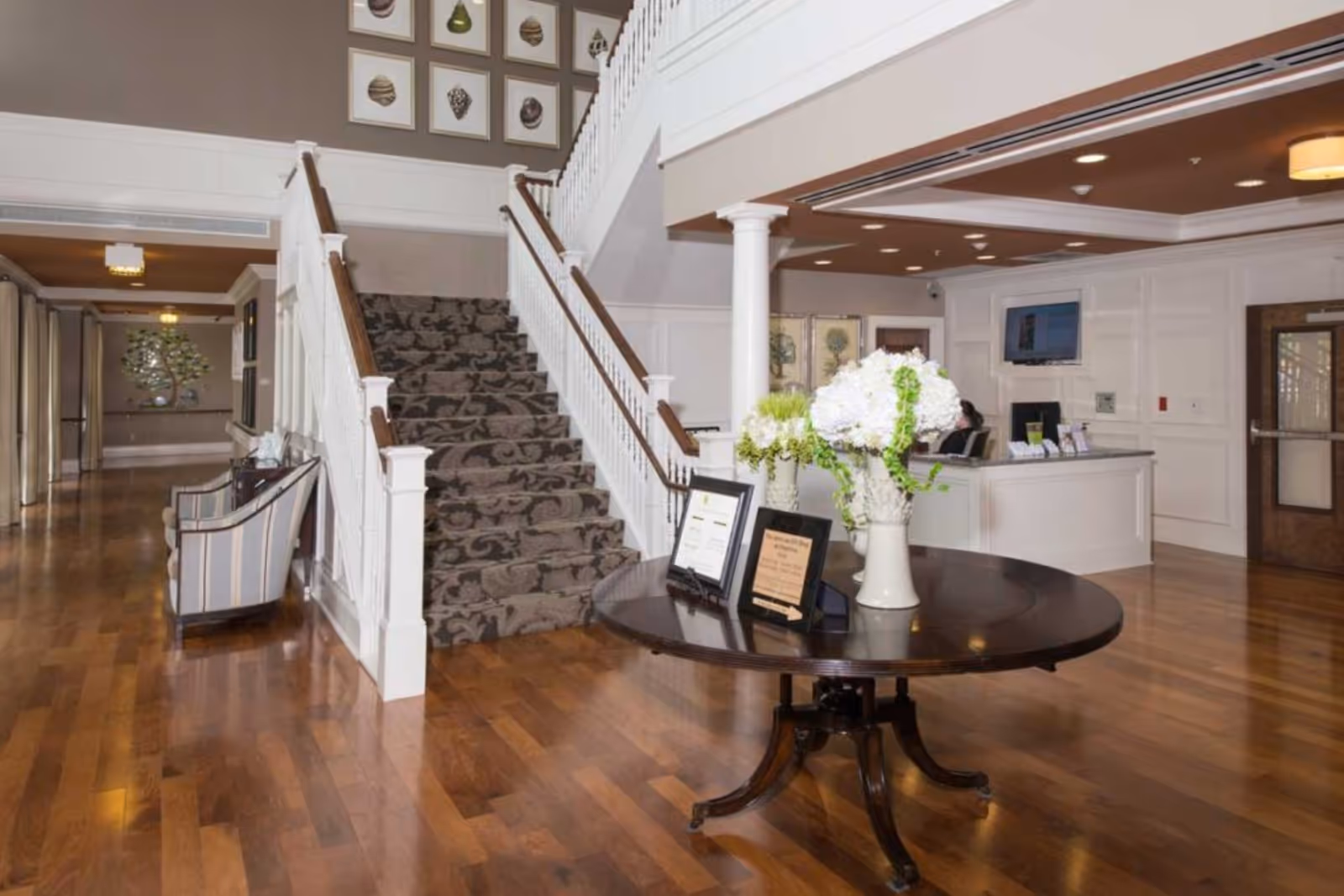 Interior view of a retirement community lobby featuring a wooden staircase with patterned carpet, a round wooden table with a vase of white flowers and framed certificates, a reception desk with a computer and a person seated behind it, and wooden flooring throughout.