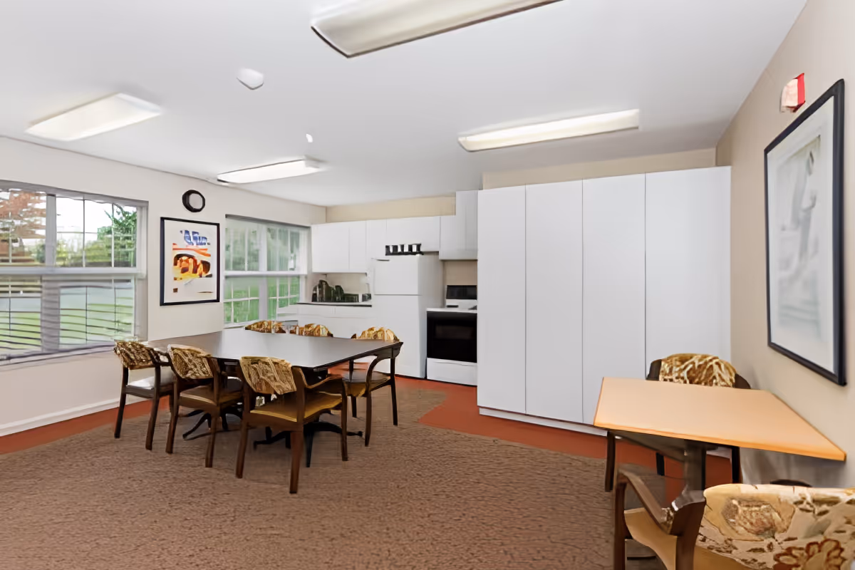 A bright room with large windows showing green outdoor scenery. Inside, there is a kitchen area with white cabinets, a refrigerator, and a stove. In front of the kitchen, there is a rectangular table surrounded by six chairs with patterned cushions. To the right, there is a smaller table with two chairs. The room has beige walls, carpeted floor, and fluorescent ceiling lights. A framed picture hangs on the wall.