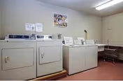 Laundry room with four white washing machines and dryers lined up against a beige wall. Above the machines are instructional signs and a framed picture. To the right, there is a folding table and a laundry cart with wheels.