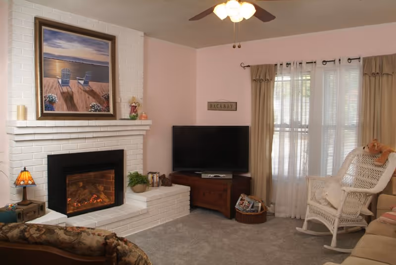 Cozy living room with a white brick fireplace, a painting above the mantel, a flat-screen TV on a wooden stand, a white wicker rocking chair with a stuffed animal, beige curtains over a window, and a ceiling fan with lights.