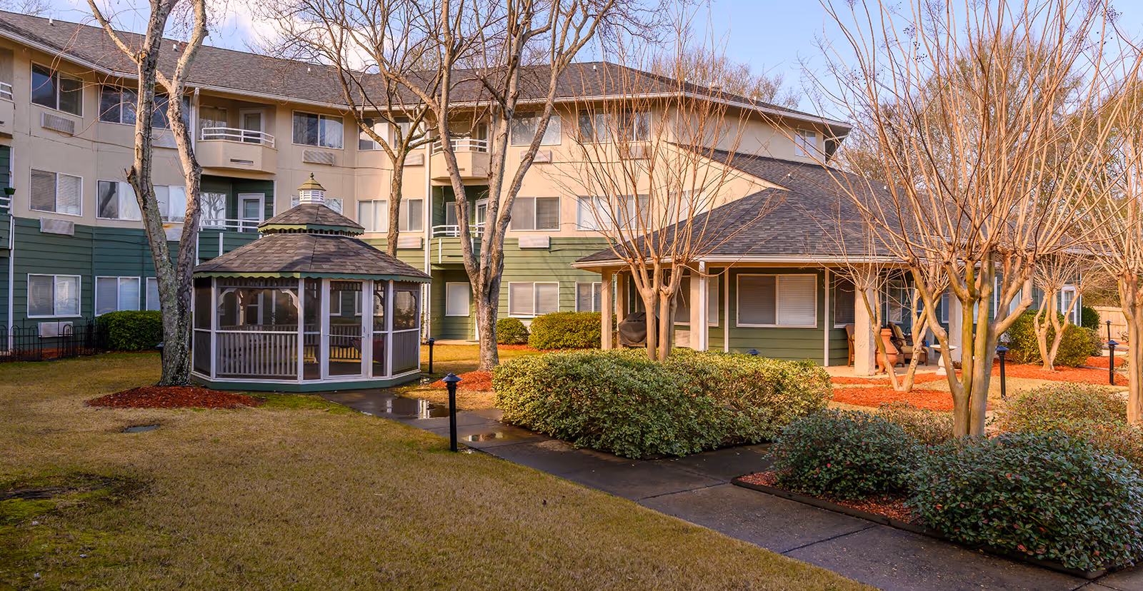 Outdoor view of Asher Point Independent Living of Montgomery featuring a multi-story residential building with green and beige exterior, a screened gazebo, leafless trees, bushes, and a paved walkway in a landscaped garden area.