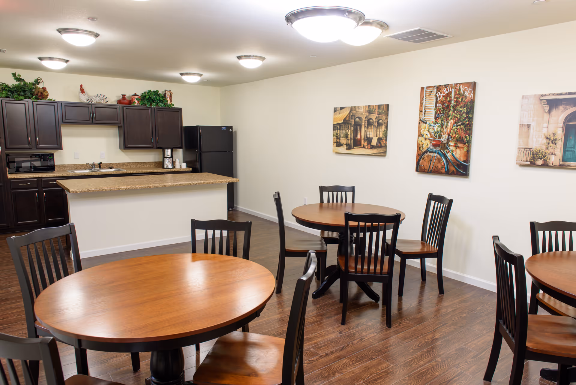 Interior view of a senior living facility dining area with round wooden tables and black chairs. In the background, there is a kitchen area with dark wood cabinets, a black refrigerator, and a countertop. The walls are decorated with framed artwork, and the floor is wooden.